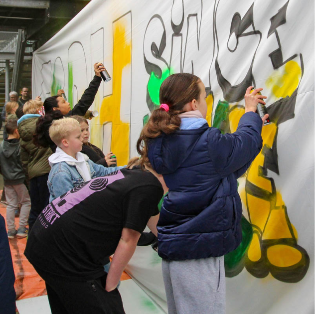 Een ADO-activiteit waar kinderen met spuitbussen aan het werk zijn om een doek te beschilderen